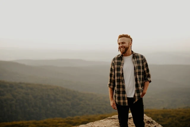Lächelnder Mann auf einem Felsgrat stehend, bewaldete Bergketten im Hintergrund